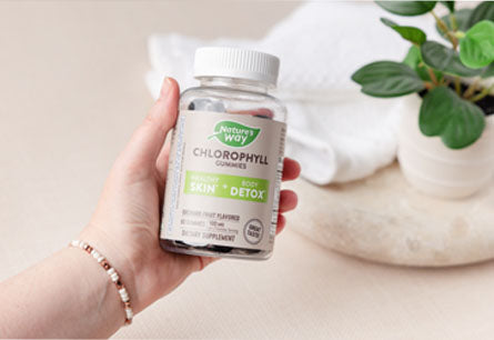A hand holding a bottle of Nature's Way Chlorophyll Gummies with a green plant and a white towel in the background.