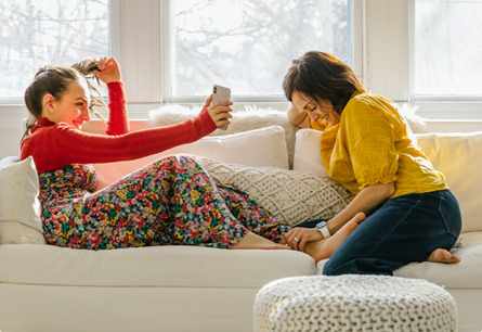 A teen girl showing her mother something on her cell phone while they're sitting and laughing together on a couch.