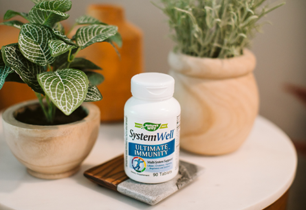 A bottle of Nature's Way SystemWell tablets on a wood and granite coaster next to some houseplants on a white side table.
