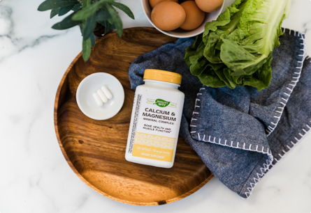 A bottle of Calcium & Magnesium laying on a tray next to a towel, green vegetables, and a bowl of eggs.