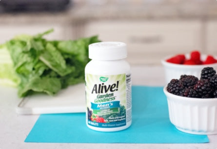 <{%DETAIL1_12112%}>A bottle of Alive Garden Goodness Men's Multivitamin sitting on a blue towel surrounded by berries and leafy green vegetables.
