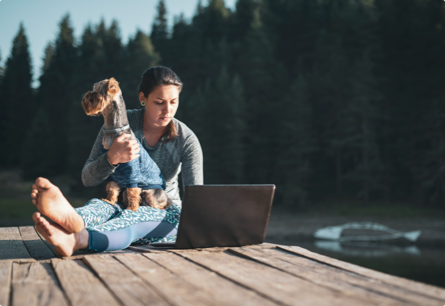 A woman sitting on a dock with a dog in her lap, she's working on a laptop.