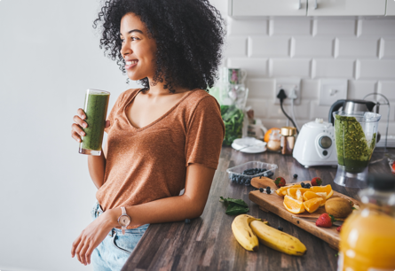 A woman holding a green smoothie leaning against a counter with a blender surrounded by fruit.