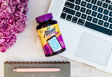 A bottle of Alive Teen Multivitamin for Her gummies laying on an open laptop next to purple flowers and a pink pen on top of a notebook.