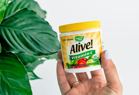 A hand holding Alive Vitamin C Powder with a green plant in the background.