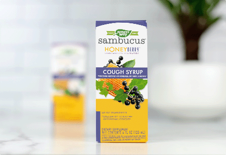 A package of Sambucus HoneyBerry Cough Syrup on a marble countertop with a second package and a plant out of focus in the background.