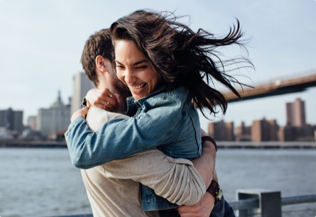 A man and a woman hugging outsided with a body of water in the background, she's laughing and her hair is blowing in the wind.