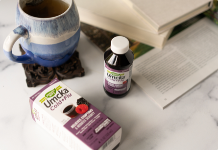 A bottle of Umcka Cold and Flu Menthol Syrup sitting on a table next to a mug, some books, and a magazine.