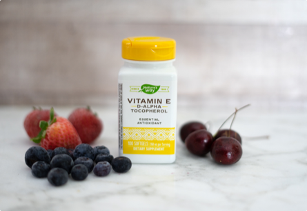 A bottle of Vitamin E on a marble counter next to blueberries, strawberries, and cherries.