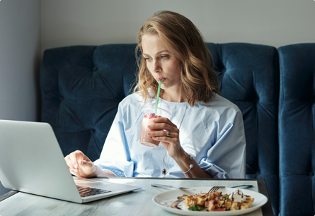 A woman sitting at a table looking at a laptop while sipping a drink next to a plate of food.