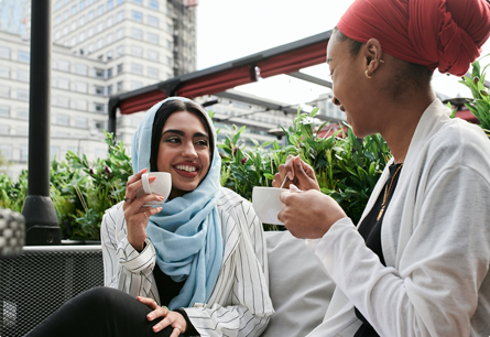 Two women sitting outside talking while holding mugs.