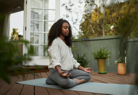A woman meditating on a wooden deck surrounded by potted plants.