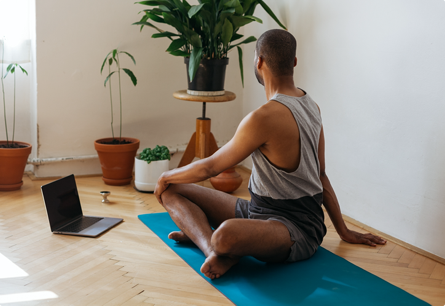 A man sitting on a yoga mat with his legs crossed, he's stretching with his back towards the camera.