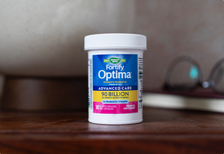 A bottle of Fortify Optima Advanced Care sitting on a dresser next to a book and a pair of glasses.
