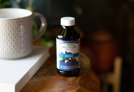 <{%DETAIL1_15803%}>A bottle of Sambucus Flu Care Syrup sitting on a wooden table top next to a coffee mug.