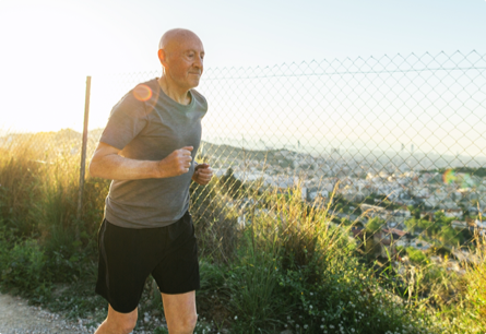 A man jogging by tall grass and a chain link fence with the sun shining behind him.