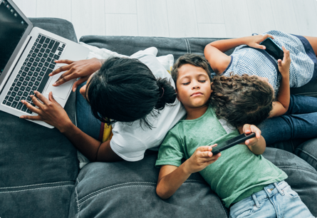 An overhead view of 3 kids leaning against each other, one is sitting up and typing on a laptop while the other two are reclined and looking at their phones.