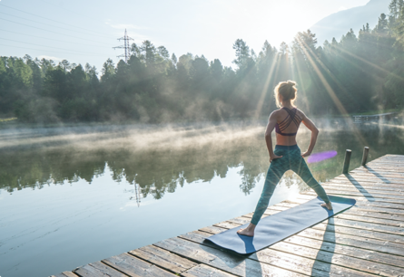 The back of a woman doing yoga on a dock with trees and the sun reflecting off of a body of water in front of her.